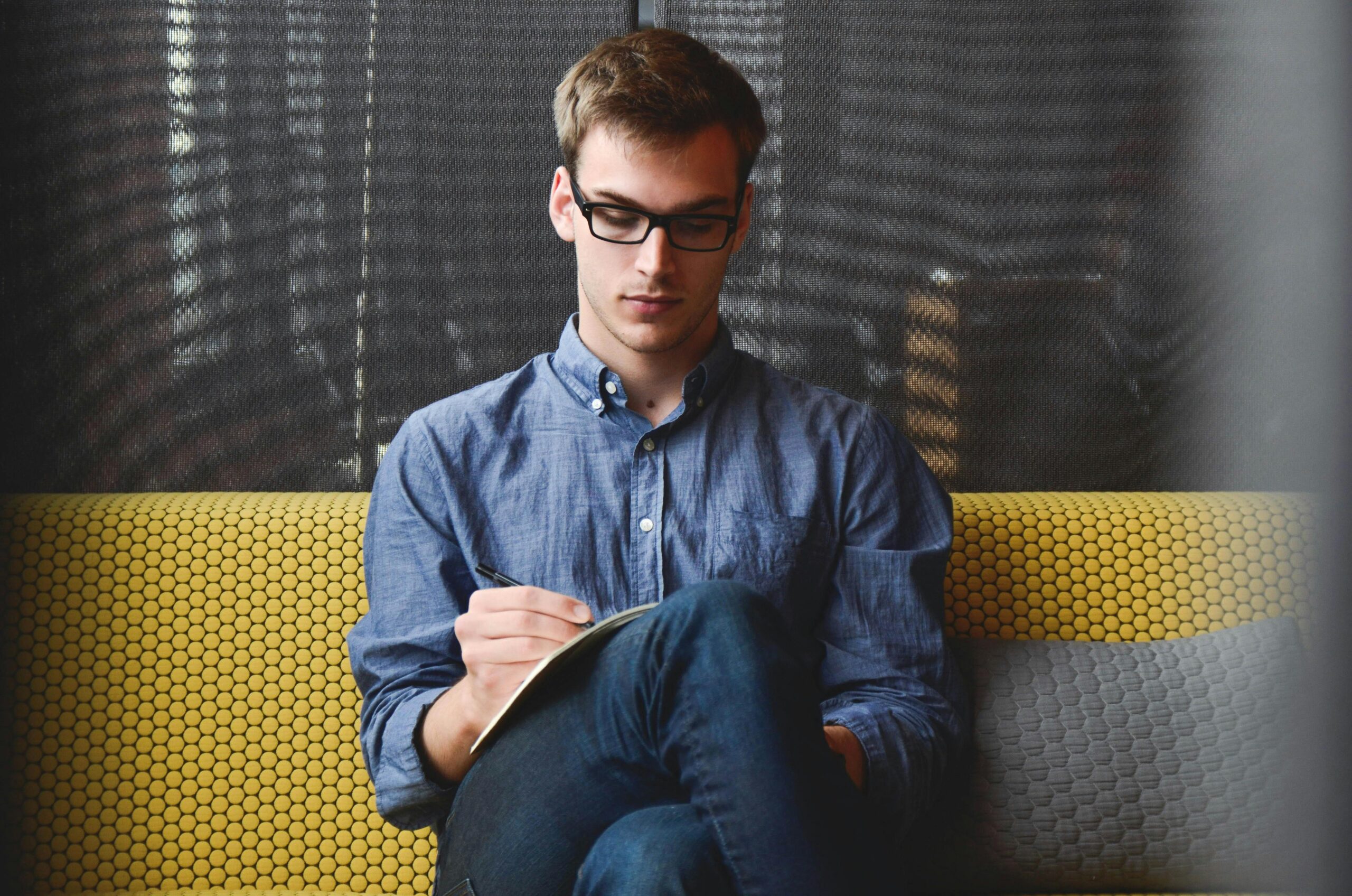 About us A young man in glasses writes in a notebook while sitting on a stylish couch indoors.